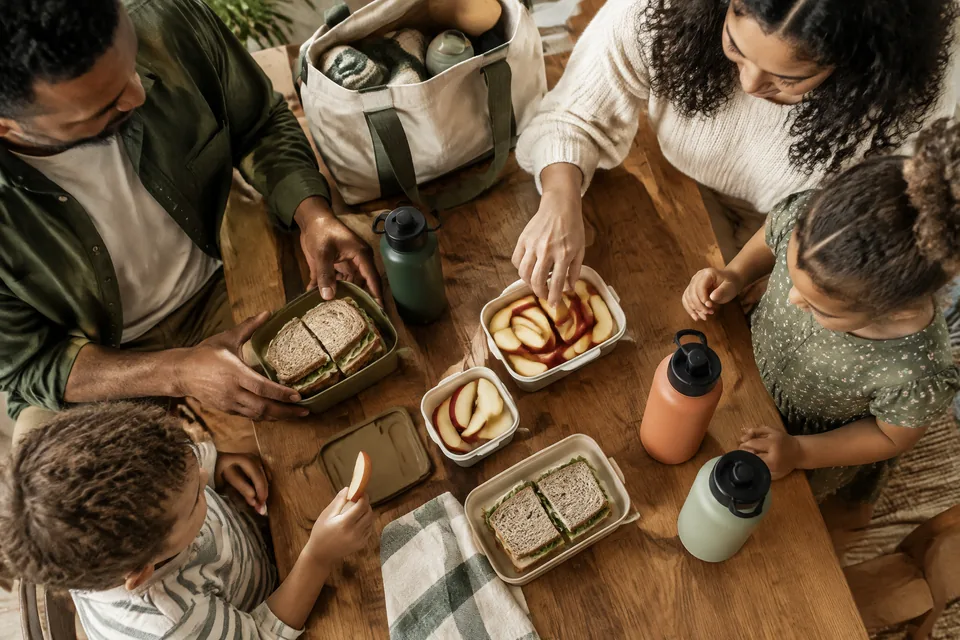 Overhead view of reusable snack containers with large apple slices, sandwiches and sturdy water bottles beside a canvas road-trip tote.